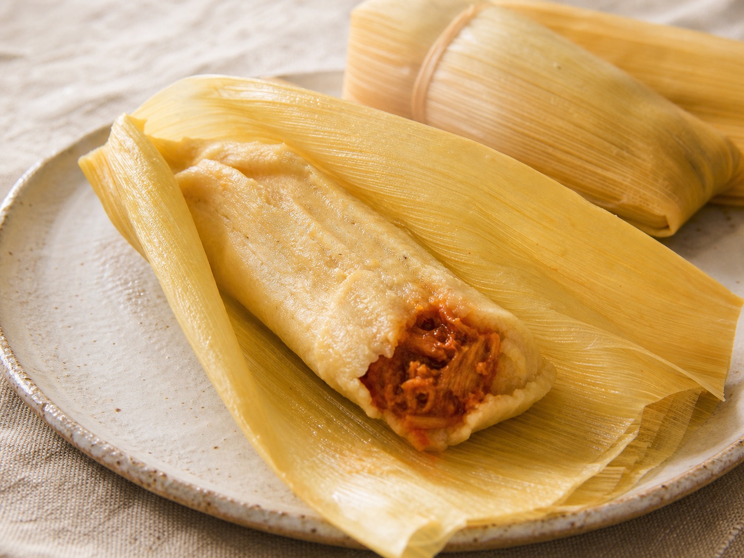 A steamed savory tamale partially unwrapped from its corn husk on a ceramic plate.
